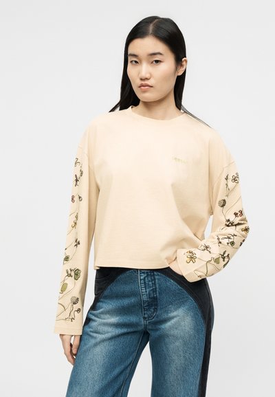 Νεαρή γυναίκα που φοράει μπεζ μπλούζα με μακριά μανίκια και κεντήματα με λουλουδένιο σχέδιο στα μανίκια, καθώς και τζιν παντελόνι δύο αποχρώσεων, μπλε και μαύρο, στέκεται με το ένα χέρι στην τσέπη.