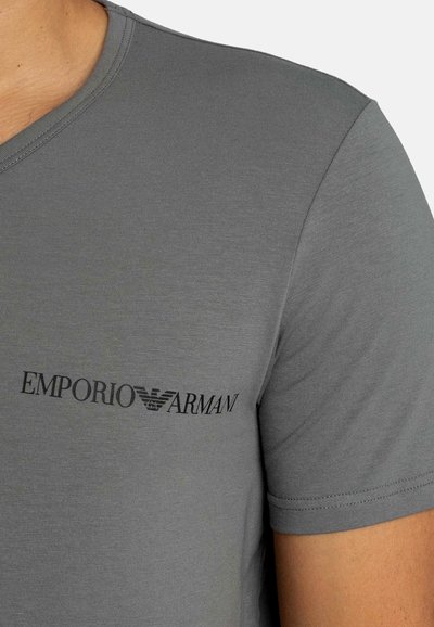 Κοντινό πλάνο ενός ατόμου που φοράει γκρι T-shirt Emporio Armani με μαύρο λογότυπο στο στήθος και κοντά μανίκια.