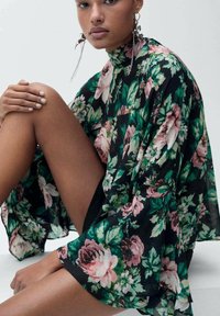 Blommig kimono med svart genomskinlig tyg, med gröna och rosa blommönster och lösa ärmar. Modellen poserar med bara ben synliga.