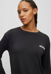 Zwarte langslavige t-shirt van zacht materiaal, met een ronde halslijn en een witte "HUGO" logo op de linkerborst.