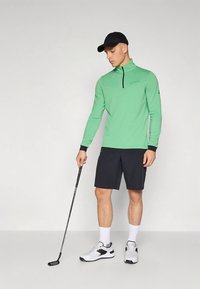 Zöld, hosszú ujjú golfpóló negyed cipzáros gallérral, fekete rövidnadrággal és fehér sportcipővel, golfütőt tartva.