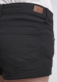 Shorts en denim noir avec un ourlet roulé, comportant deux poches arrière et une étiquette en cuir marron sur la taille marquée "TWIN TIP."