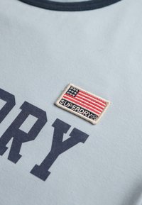 Hellblaues Baumwoll-T-Shirt mit einem marineblauen Rundhalsausschnitt, fettem "SUPERDRY"-Schriftzug in Dunkelblau und einem aufgestickten amerikanischen Flaggen-Patch.