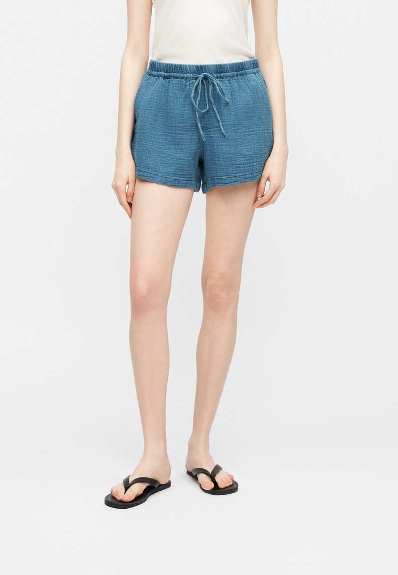 Personne portant un short bleu texturé avec cordon de serrage, un haut blanc sans manches et des tongs noires, debout sur un fond clair uni.