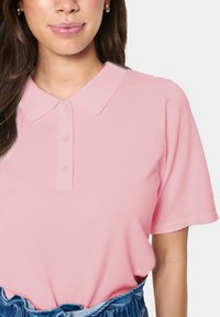 Polo-paita pehmeästä vaaleanpunaisesta ribbimateriaalista, jossa on kaulus, kolme nappia kauluksessa ja lyhyet hihat, yhdistettynä farkkutukeen.