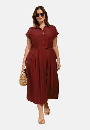 Femeie purtând o rochie midi burgundy cu nasturi și curea, sandale aurii, ochelari de soare, ținând o geantă țesută, stând în fața unui fundal simplu.