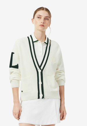 Femme portant un cardigan blanc avec des rayures vert foncé sur un polo blanc et une jupe blanche, debout devant un fond uni.