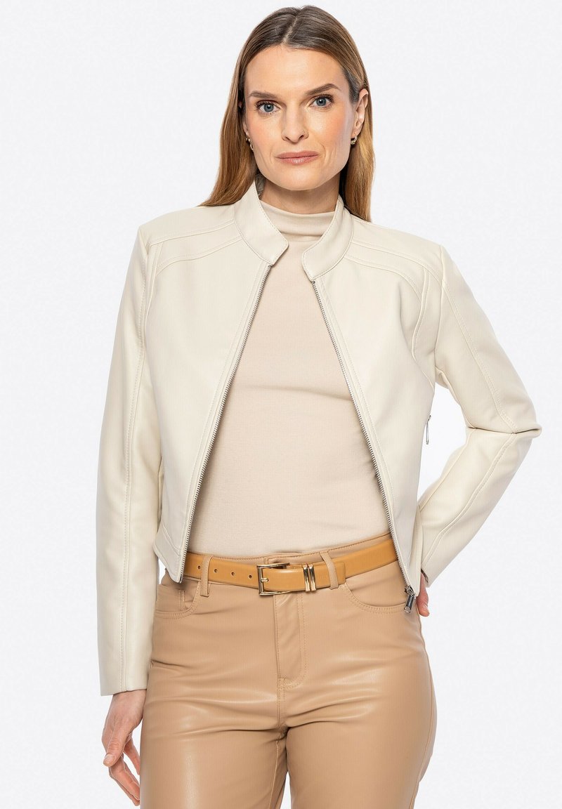 Giacca di pelle crema con colletto alto, design aderente e dettagli di cucitura sottili. Indossata sopra una maglietta beige e pantaloni marrone chiaro con una cintura.