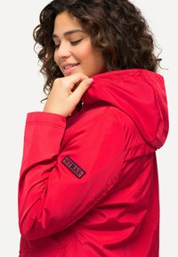 Giacca impermeabile rossa con cappuccio, caratterizzata da una patch logo nera sulla manica, superficie liscia e chiusura frontale con zip.