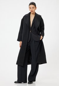 Cappotto in lana nera con ampio colletto, vestibilità over size, tasche frontali e chiusura con un solo bottone su un outfit elegante nero.