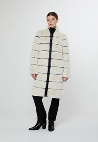 Kabát z umělé kožešiny v krémové barvě s černými vodorovnými pruhy, s vysokým límcem, plným předním zipem a bočními kapsami.