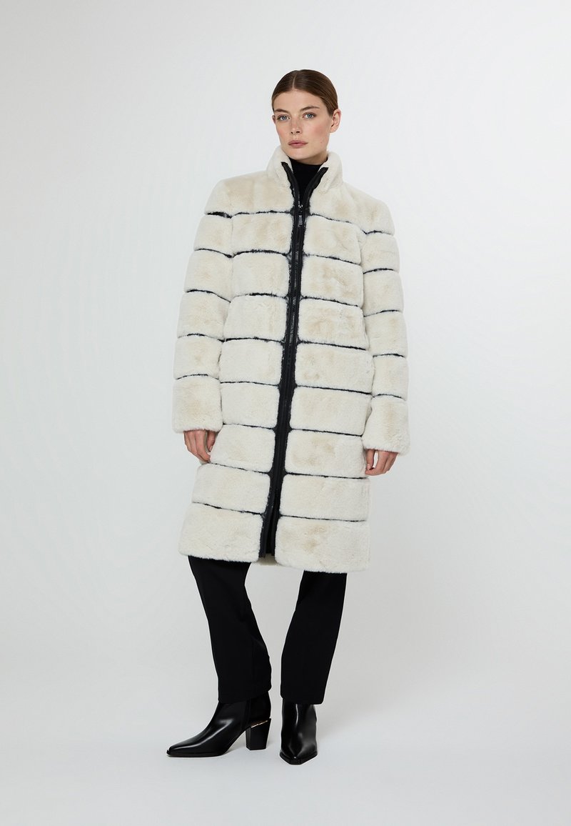 Kabát z umělé kožešiny v krémové barvě s černými vodorovnými pruhy, s vysokým límcem, plným předním zipem a bočními kapsami.