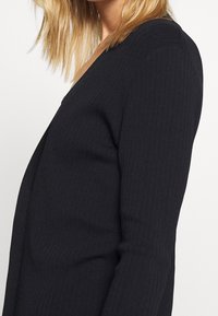 Schwarze gerippte Strickjacke mit tiefem V-Ausschnitt und langen Ärmeln. Die weiche Textur und die taillierte Silhouette betonen ihr strukturiertes Design.