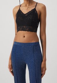 Bralette en dentelle noire avec des bretelles ajustables, présentant un ourlet festonné et des motifs floraux complexes, associé à un pantalon fuselé bleu scintillant.