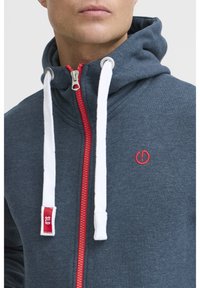 Uomo che indossa un hoodie blu scuro con zip rossa, lacci bianchi e un piccolo logo circolare rosso sul petto.