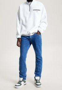 Pullover in pile bianco con collo a zip, abbinato a jeans in denim blu e sneakers grigie/bianco con accenti neri e suola piatta.