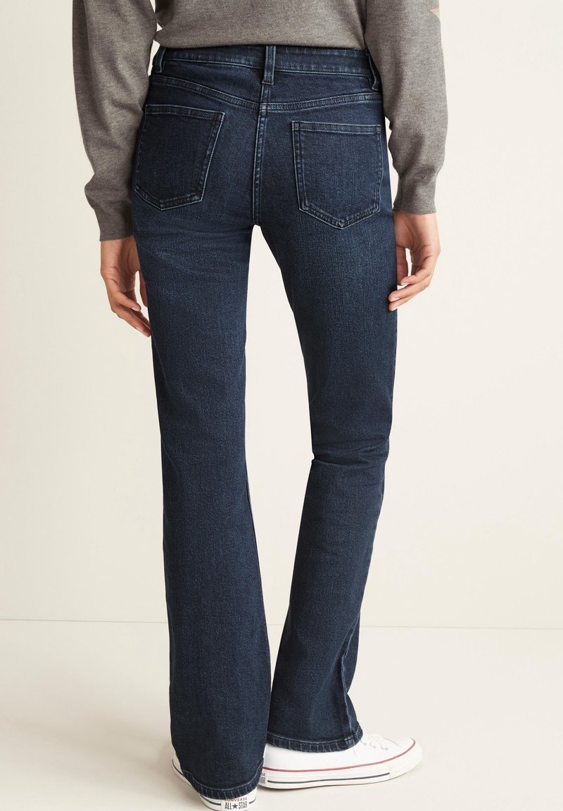 Personne portant un jean bootcut bleu foncé, un haut gris à manches longues et des baskets blanches, debout de dos sur un fond uni.