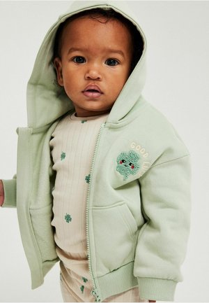 Junges Kind trägt eine hellgrüne Kapuzenjacke mit der Aufschrift "Viel Glück" und einem Kleeblatt-Aufnäher sowie ein beiges Outfit mit kleinen grünen Blumenmustern.