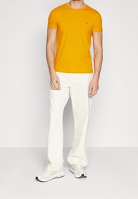 Man som bär en figursydd kortärmad orange t-shirt, vita lösa byxor och vita sneakers som står mot en neutral ljus bakgrund.