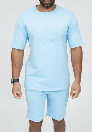 Homme portant une chemise à manches courtes bleu clair et un short assorti avec un logo "ICON ORIGINAL" en relief sur la poitrine et une montre connectée au poignet gauche.