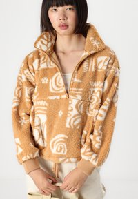 Fleece-Pullover in Beige mit weißen abstrakten Mustern, ausgestattet mit einem halb hohen Reißverschluss-Kragen, elastischen Bündchen und einer lockeren Passform. Glatte Innenstruktur.