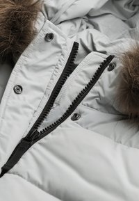 Lys grå pufferjakke med en sort lynlås foran, trykknapper og en faux fur hættekant, der har quiltet tekstur og mat finish.