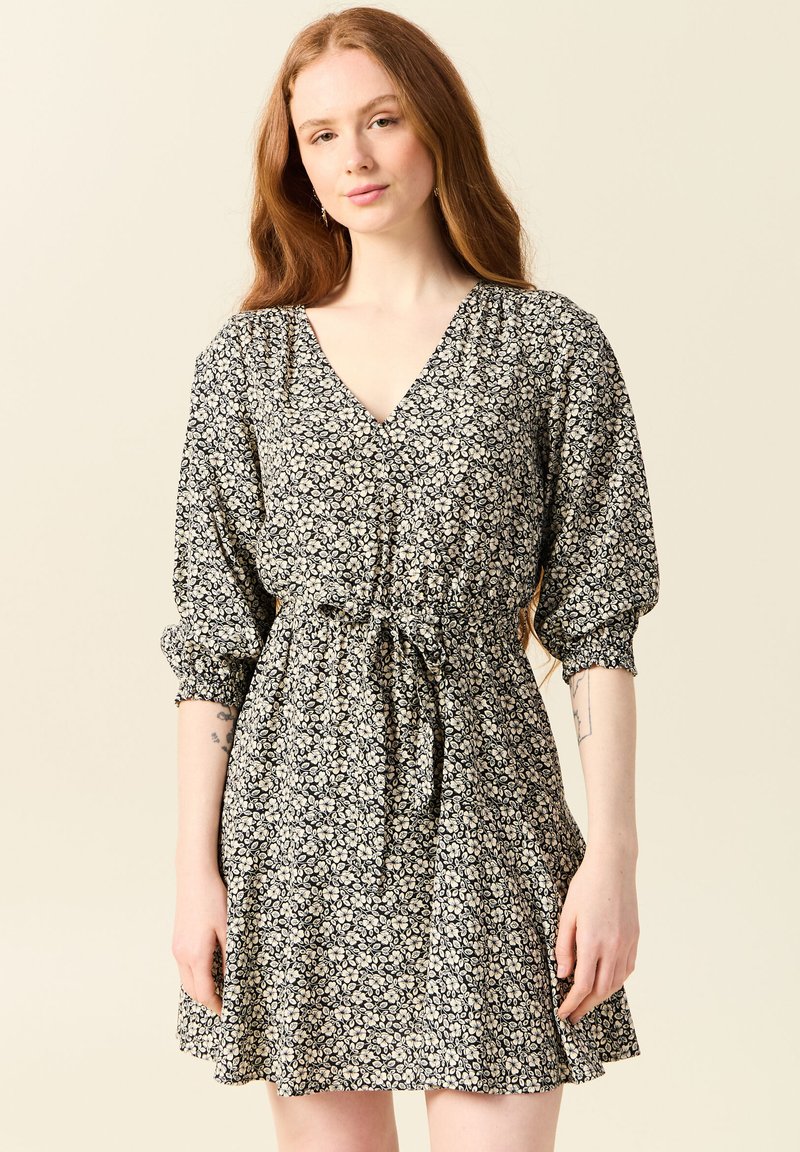 Svart blommig klänning med v-ringning, puffärmar, rynkad midja och knytbälte. Har ett mönster av små vita blommor på en mörk bakgrund.