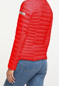 Červená prošívaná bunda s prošívaným vzorem. Má kulatý límec, boční kapsy a hladkou látku. Prošití zdůrazňuje konstrukci.