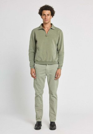 Modèle masculin debout, portant un sweat-shirt à col zippé vert clair, un pantalon slim assorti et des mocassins noirs, sur fond blanc uni.