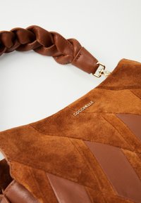Brown suede and leather handbag with a braided strap, featuring geometric patterns and a metallic logo detail.