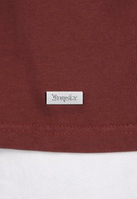 Mørkerød bomulds t-shirt med en glat tekstur, der har et sølvfarvet rektangulært mærke med teksten "Dropsize" nær sømmen.