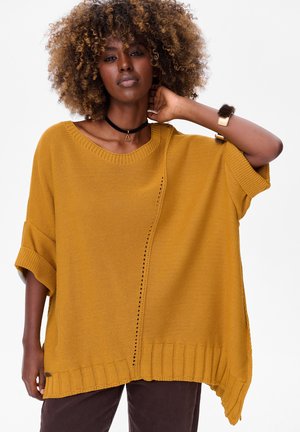 Kobieta o kręconych włosach, ubrana w musztardowy, oversize'owy sweter z dzianiny i czarny choker, pozująca z jedną ręką na szyi na białym tle.