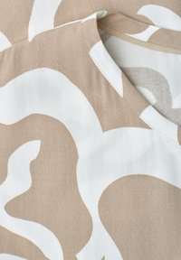Gros plan sur un tissu beige et blanc à motifs avec des formes courbes et des coutures visibles au niveau de l'encolure d'un vêtement.