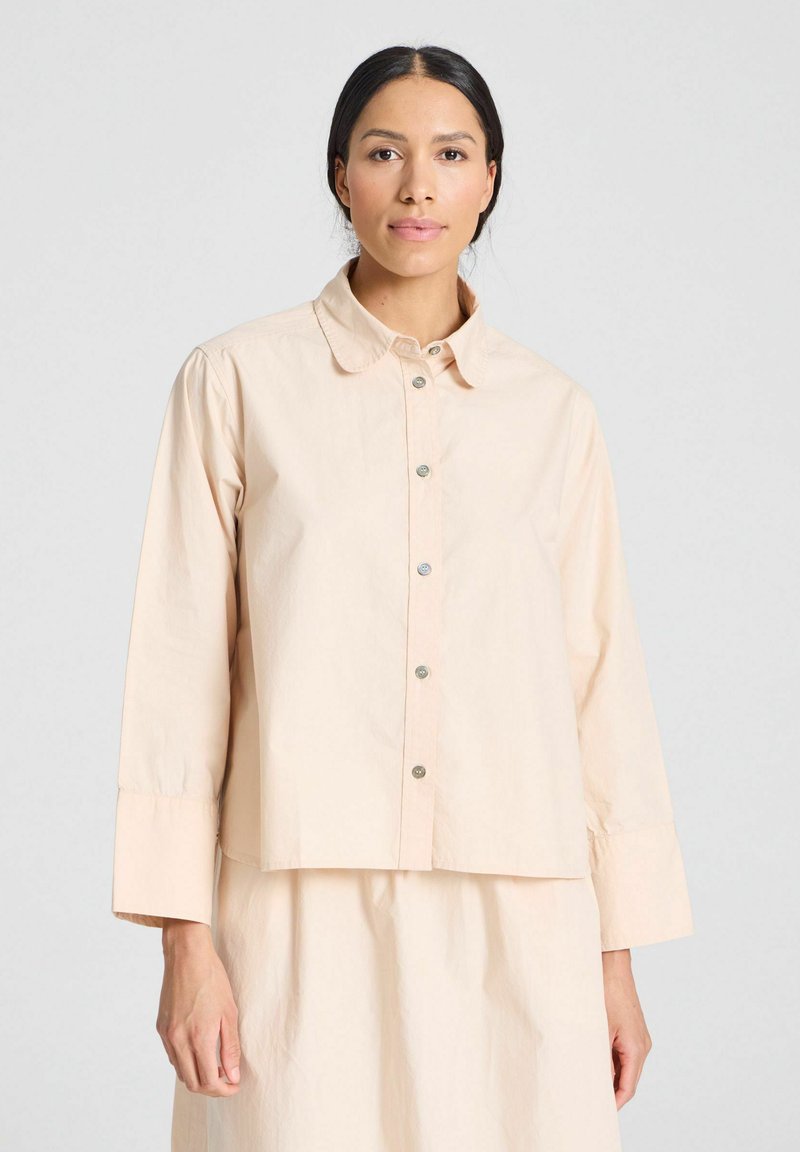 Camisa de botones de manga corta en un suave color durazno claro, confeccionada con una tela lisa. Presenta un cuello clásico y botones a presión plateados.