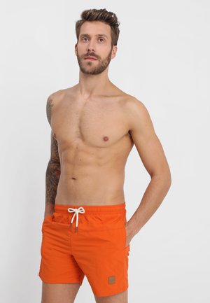 Oranžové plavky vyrobené z ľahkého materiálu s pásom na stiahnutie a malou logo nášivkou na ľavom stehne. Hladká textúra, dĺžka nad kolená.
