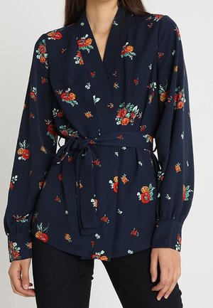 Blouse portefeuille bleu marine à motifs floraux avec manches longues et ceinture à nouer, portée avec un pantalon noir.