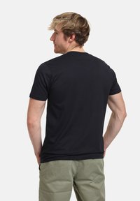 Černé tričko s krátkým rukávem a kulatým výstřihem. Hladká textura a přiléhavý střih, zobrazeno na modelce zezadu, v kombinaci se světlými zelenými kalhotami.