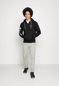 Felpa nera con zip e logo bianco sul lato sinistro del petto e sulla manica, abbinata a pantaloni della tuta grigio chiaro e sneakers nere con suole color gomma.