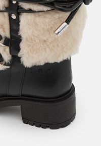 Botte en cuir noir avec semelle épaisse et doublure en laine de mouton de couleur crème, équipée d'œillets métalliques et d'un cordon de serrage avec embout métallique.