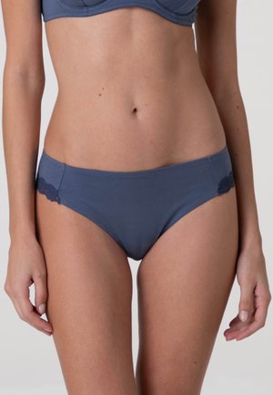 Donna che indossa biancheria intima blu in stile hipster con dettagli in pizzo ai lati, mostrata dal torso medio alla parte superiore delle cosce su uno sfondo semplice.