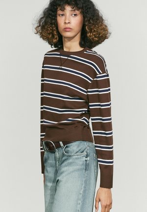 Person mit lockigem Haar, die ein braun gestreiftes Langarmshirt und helle blaue Jeans trägt, steht vor einem einfarbigen Hintergrund.