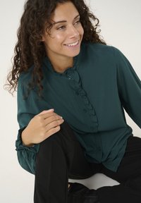 Blusa ruffled di un verde scuro con bottoni frontali e maniche lunghe con polsini arricciati, abbinata a pantaloni neri. Tessuto a trama liscia.