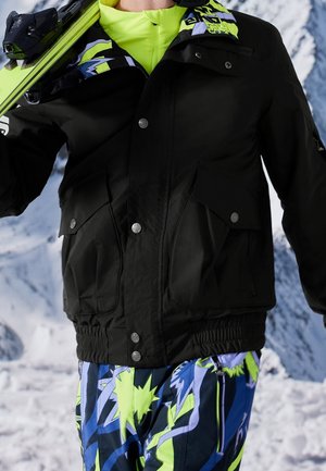 Personne en veste noire et pantalon de ski coloré portant des skis vert néon sur un fond de montagne enneigée.