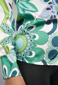 Saténová halenka s výrazným zeleno-modrým květinovým vzorem a kulatým, detailním středem. Obsahuje kontrastní manžetu a hladkou texturu.