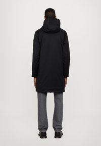 Must must pikk parka kapuutsiga, valmistatud siledast materjalist. Omab sirget lõiget ja on koos hallide pükste ning mustade kingadega.