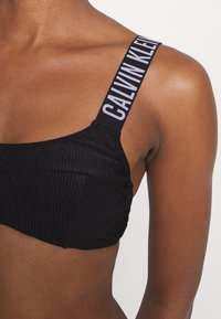 Črn rebrast bralette z širokimi blagovnimi trakovi, ki nosijo napis "CALVIN KLEIN", ima gladko teksturo in strukturiran dizajn.