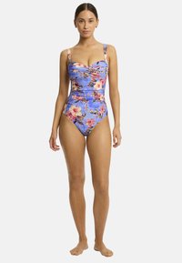 Model in een blauw badpak met bloemmotief staat blootsvoets tegen een effen achtergrond, waarbij het ontwerp en de pasvorm van het kledingstuk worden getoond.