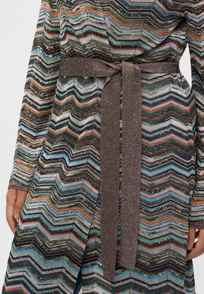 Femme portant une robe en maille à manches longues avec des motifs zigzag multicolores et une ceinture métallique marron nouée à la taille.