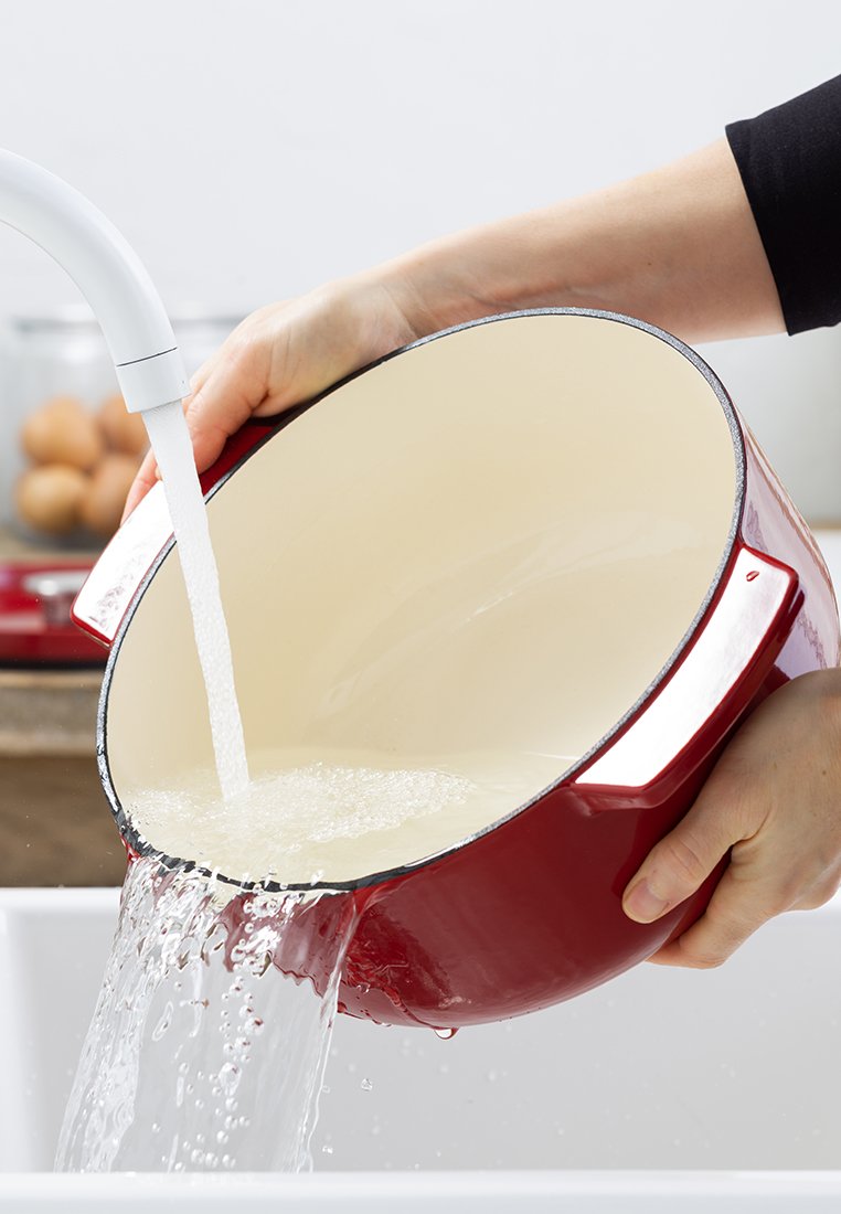 Roter Emaille-Topf mit glattem, crema-farbenem Innenraum. Wasser wird aus dem Topf unter einem Wasserhahn gegossen. Der Topf verfügt über zwei robuste Griffe.