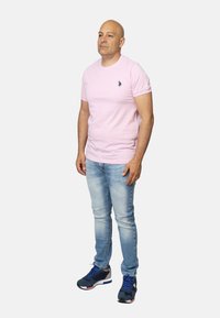 Maglietta rosa a maniche corte con un piccolo emblema, abbinata a jeans blu chiaro e scarpe da ginnastica nere con accenti rossi e blu.